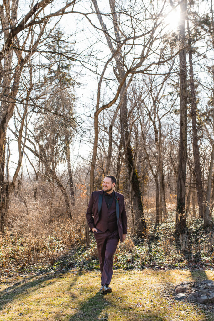 Groom walking by woods with hand in his pocket.