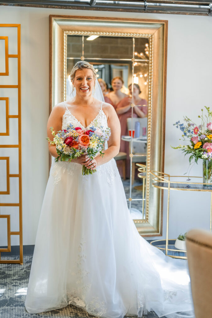 Bride smiles in front of mirror with reflections of bridesmaids.