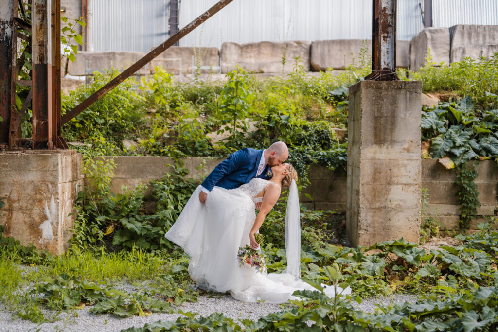 Bride and groom share a dip kiss in front of a vine-covered industrial area at One World at Woolery Mill.