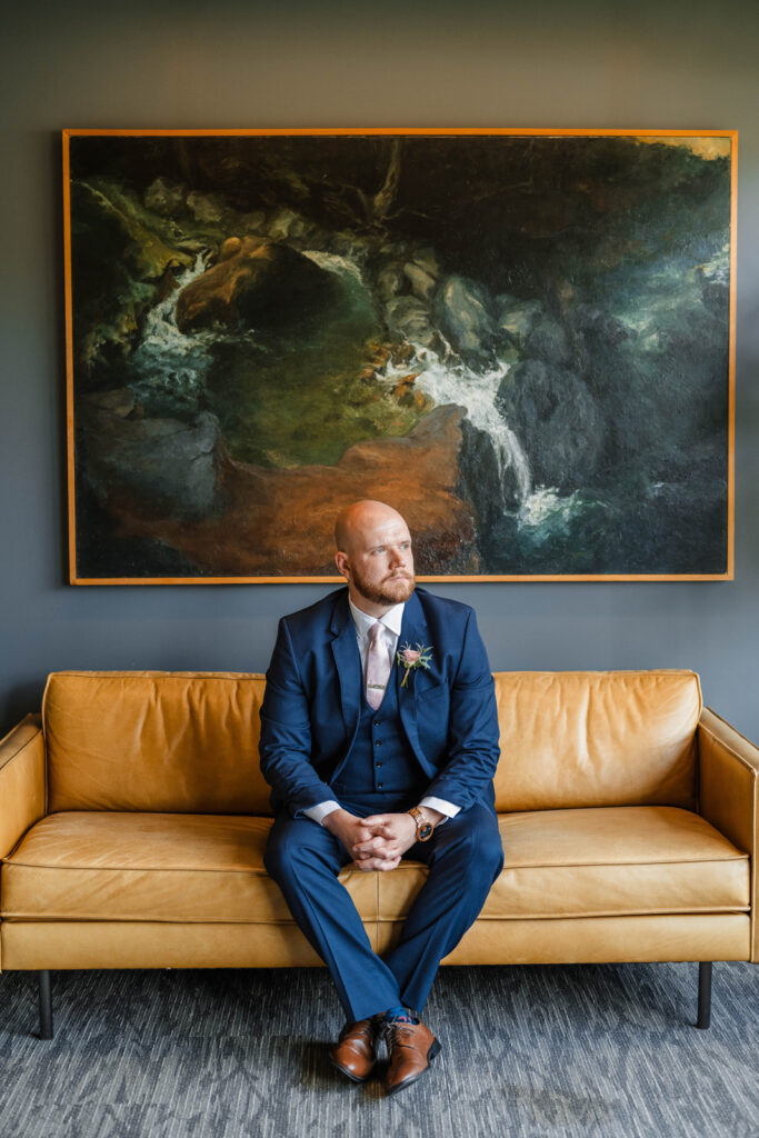 Groom sits on yellow couch looking out towards window.