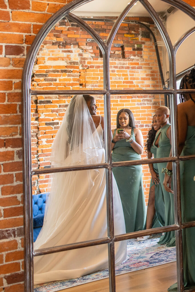 Bride surrounded by bridesmaids looks at her reflection in arch mirror.