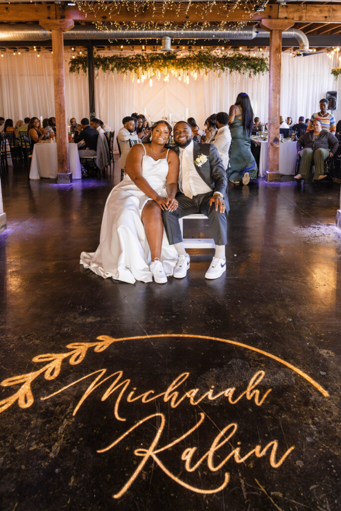 Bride and groom sitting at chairs during their reception smiling in front of custom sign at Ivory Foundry Weddings & Events.