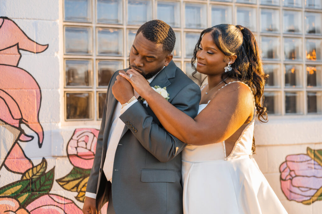 Groom kisses bride's hand while standing in front of mural at Ivory Foundry Weddings & Events.
