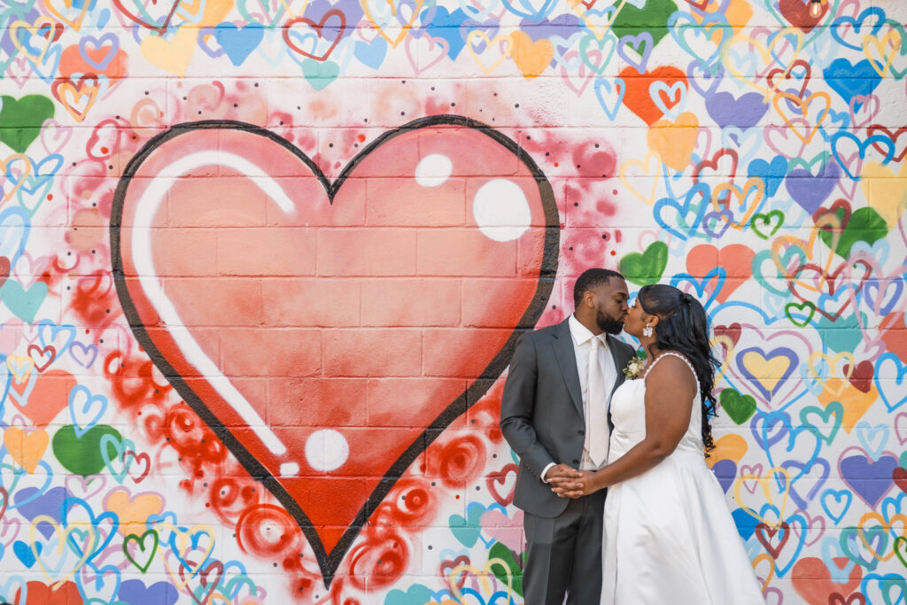 Couple kiss in front of a heart mural on the side of a building at Ivory Foundry Weddings & Events.