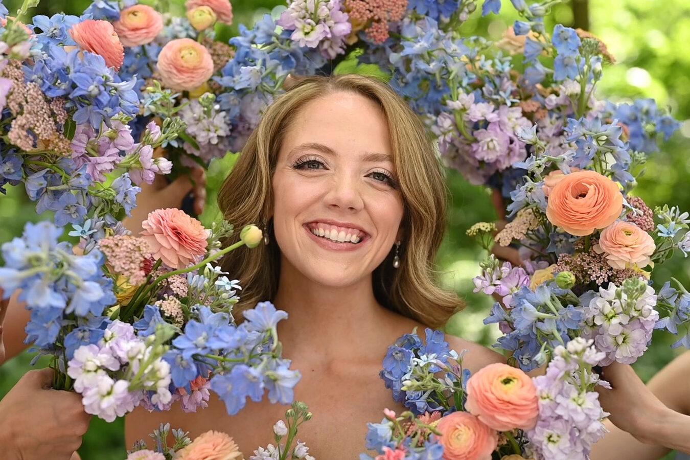 Bride smiling while bridesmaids hold flower bouquets around her face.