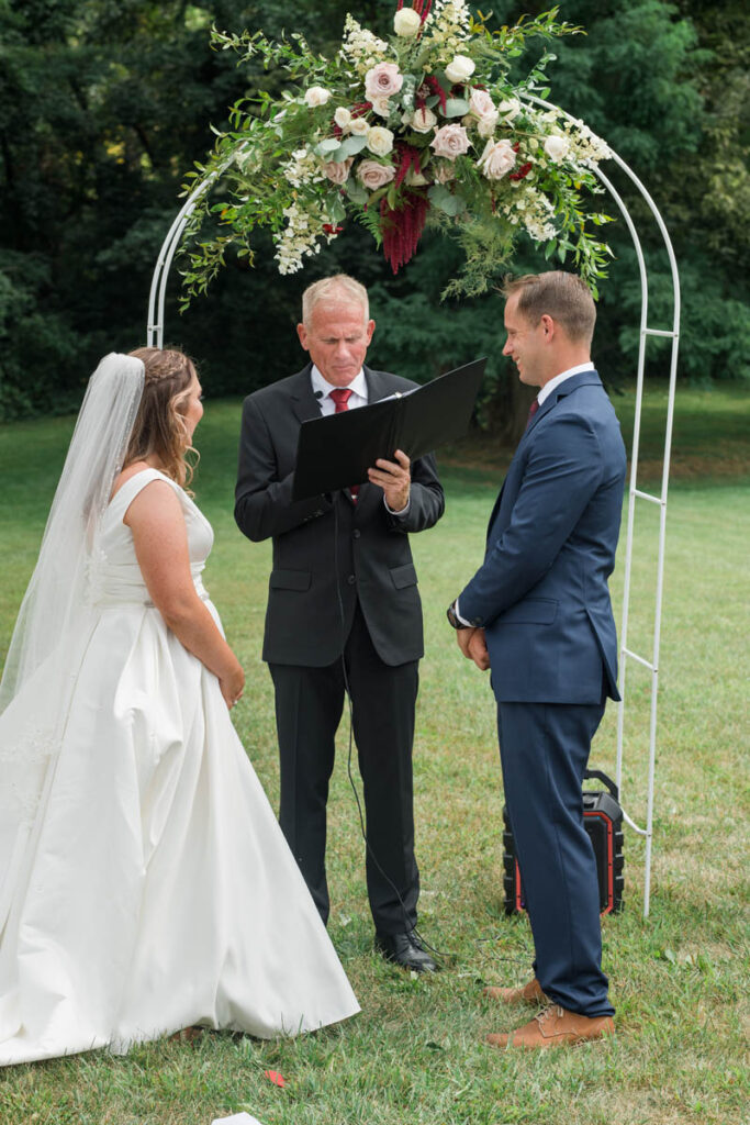 Officiant marries bride and groom under a floral arch at an Indiana outdoor wedding.