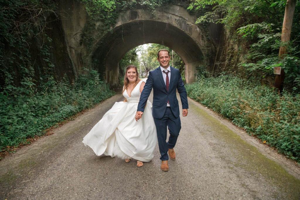 Bride and groom hold hands while walking away from an old bridge.