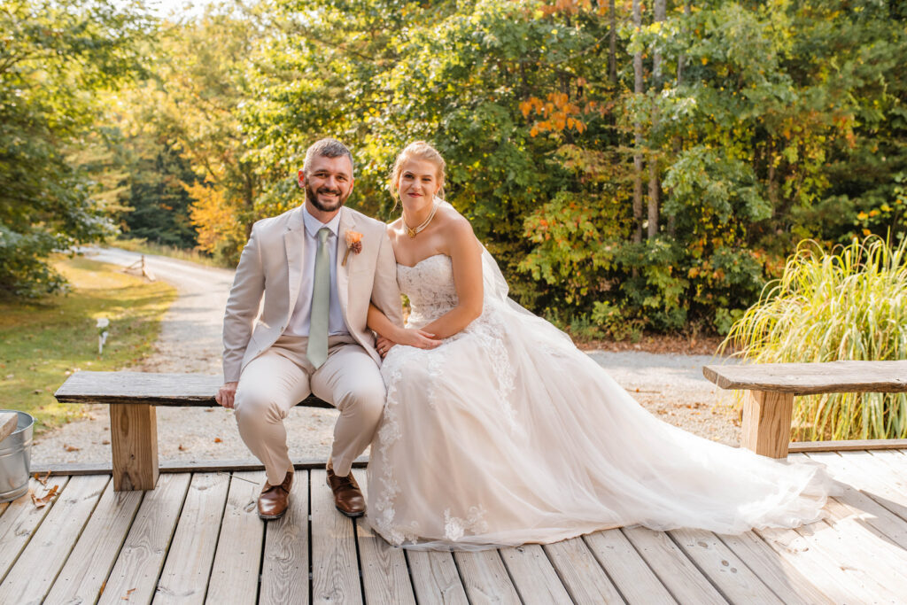 Bride and groom sit together on a bench surrounded by the woods.