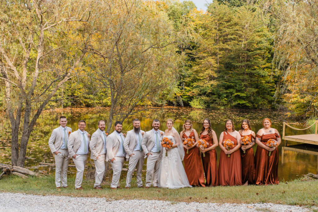 Wedding party stands by a pond, dock, and trees in the autumn at The Barn at Timber Ridge.